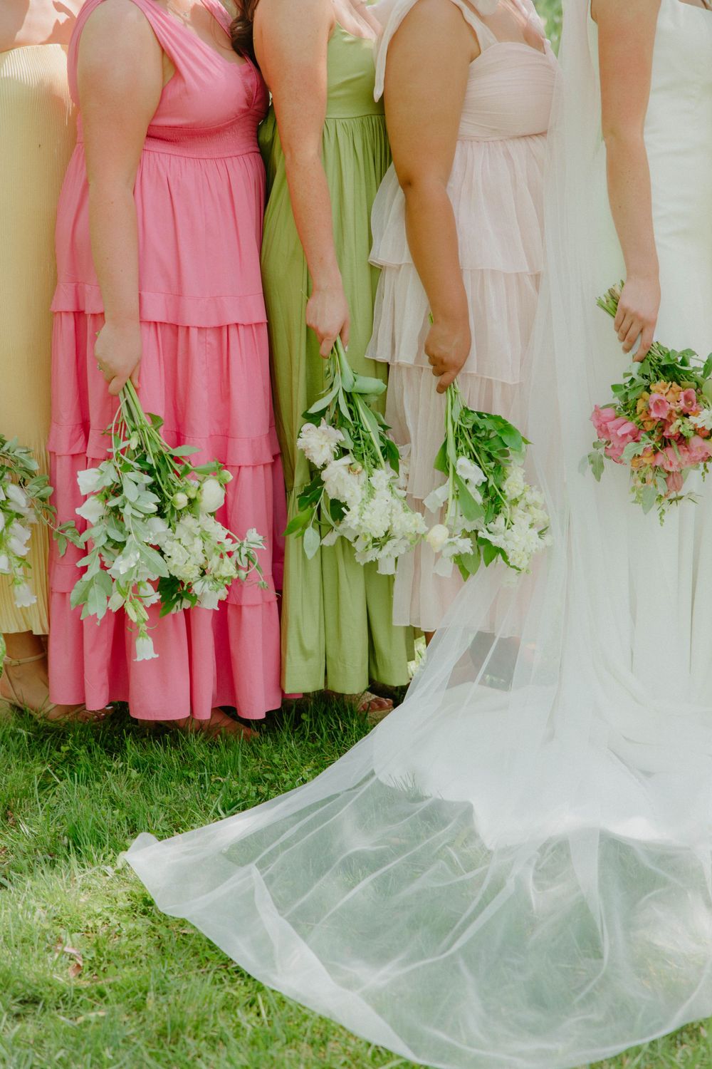 Bridesmaids in pink and mint dresses holding fresh flower bouquets stand beside a wedding gown train.