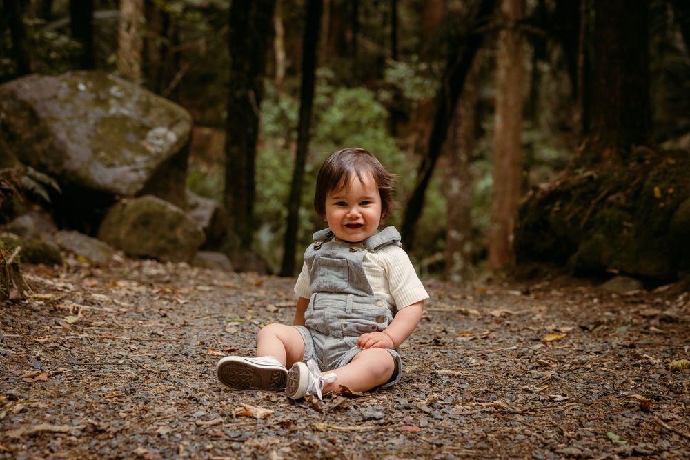 A small child sits on a forest path wearing a grey outfit surrounded by woodland scenery.