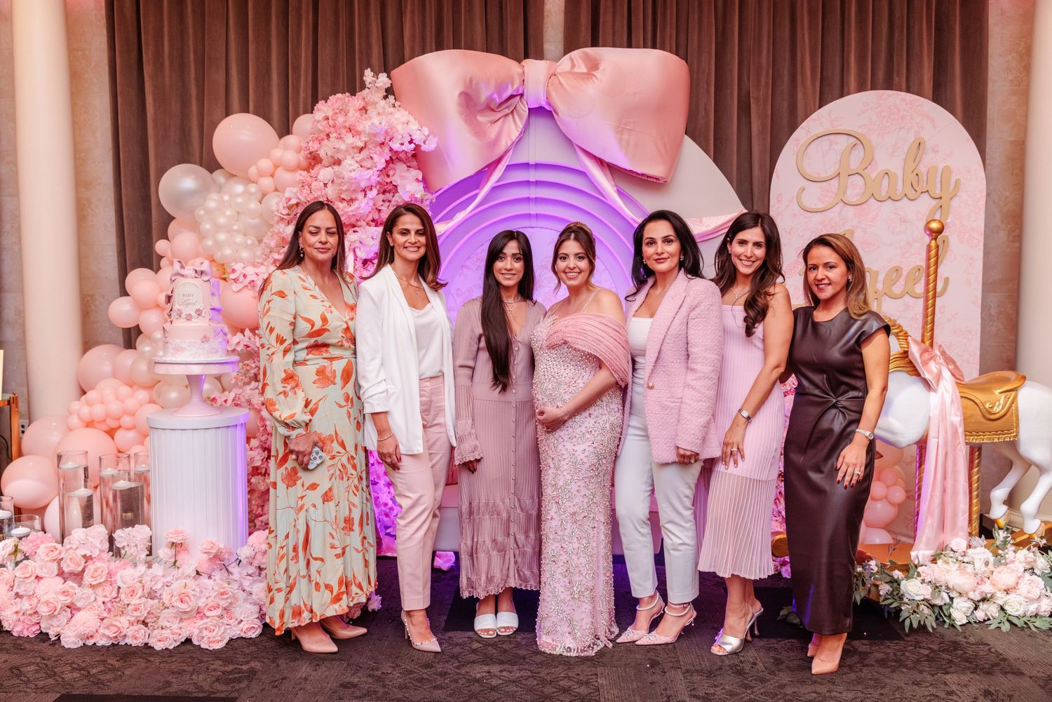 A baby shower celebration photo featuring pink and white decor with guests in formal attire at an elegant venue.