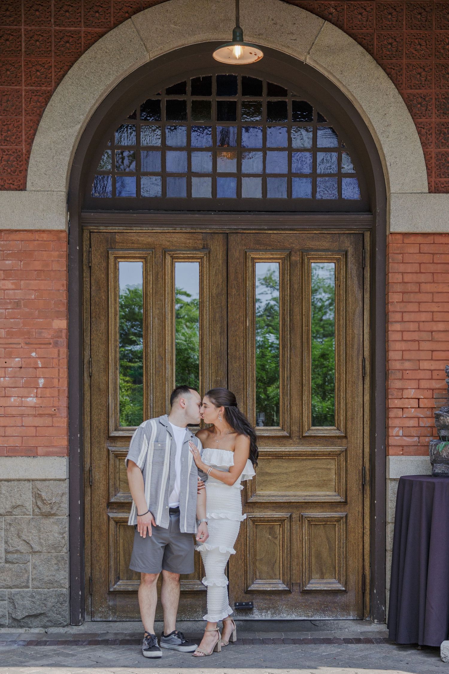 A couple stands together in front of a grand wooden arched doorway with vintage brick and stonework details.