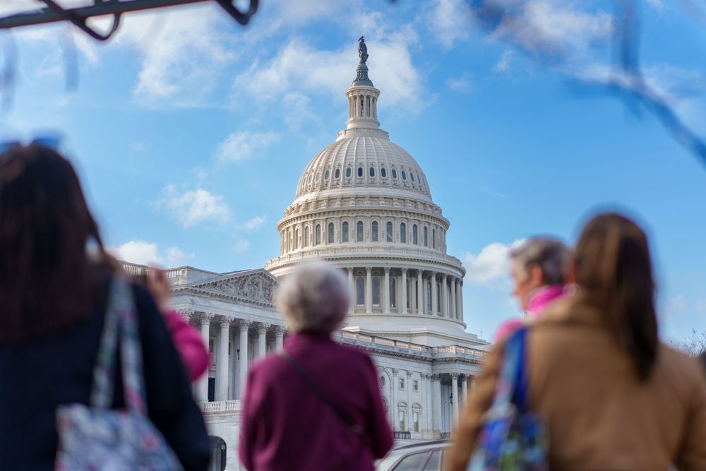 A diverse group of people view the iconic US Capitol Building dome on a sunny day against a blue sky.