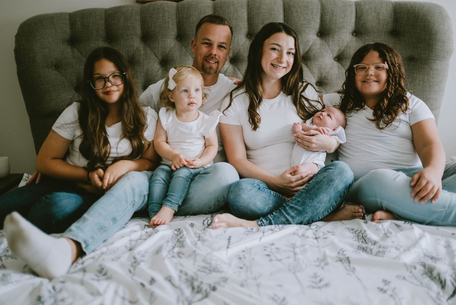 Family members lounging together on a cozy white bed wearing casual denim and neutral colors.