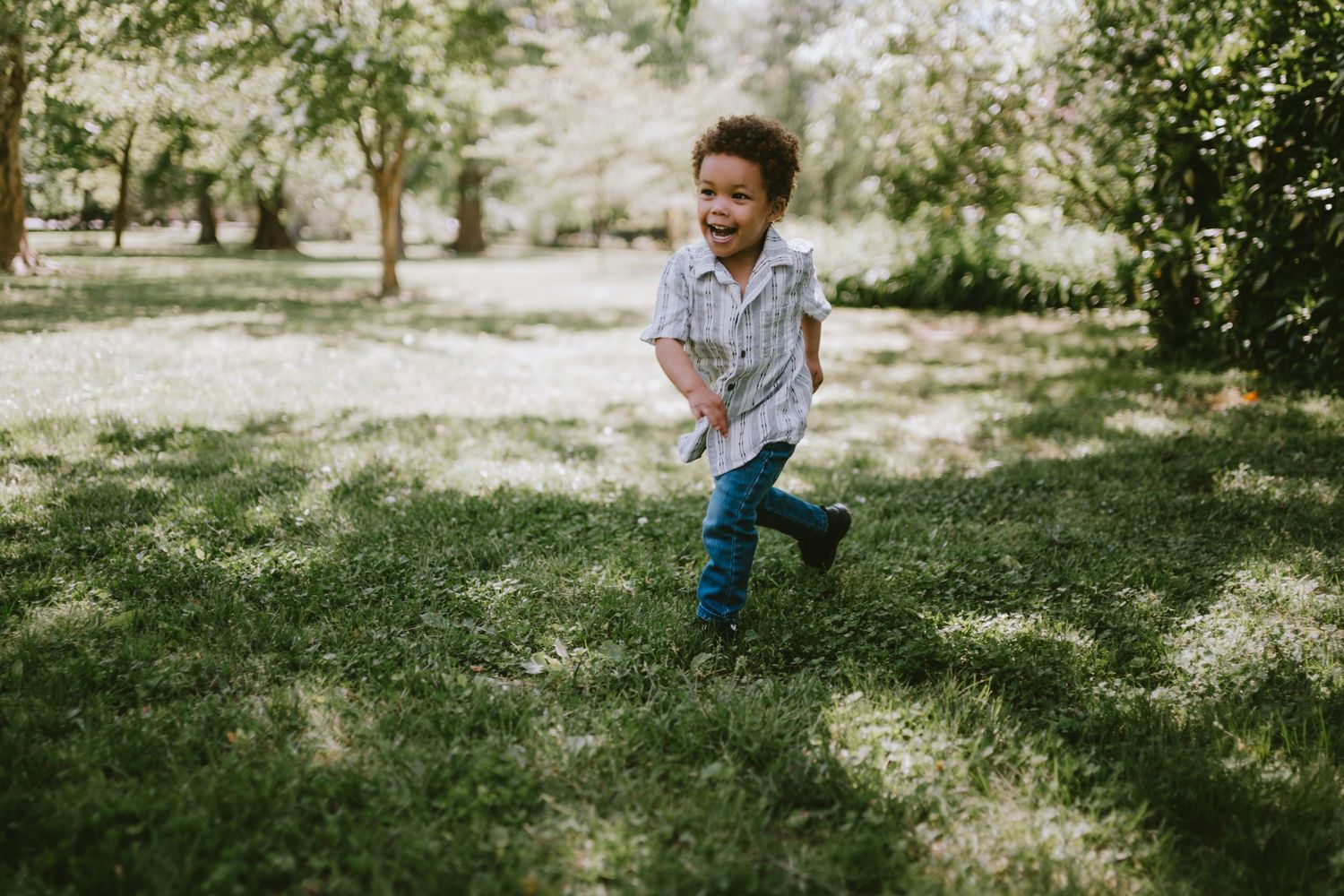 Happy young boy running and playing outdoors on lush green grass in a sunny park surrounded by trees.