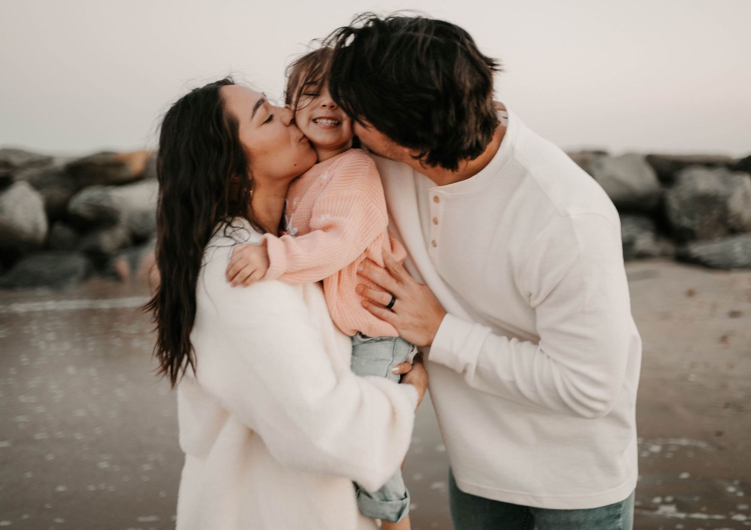 Parents kissing their smiling toddler on the cheeks at a rocky beach during a family photo session.