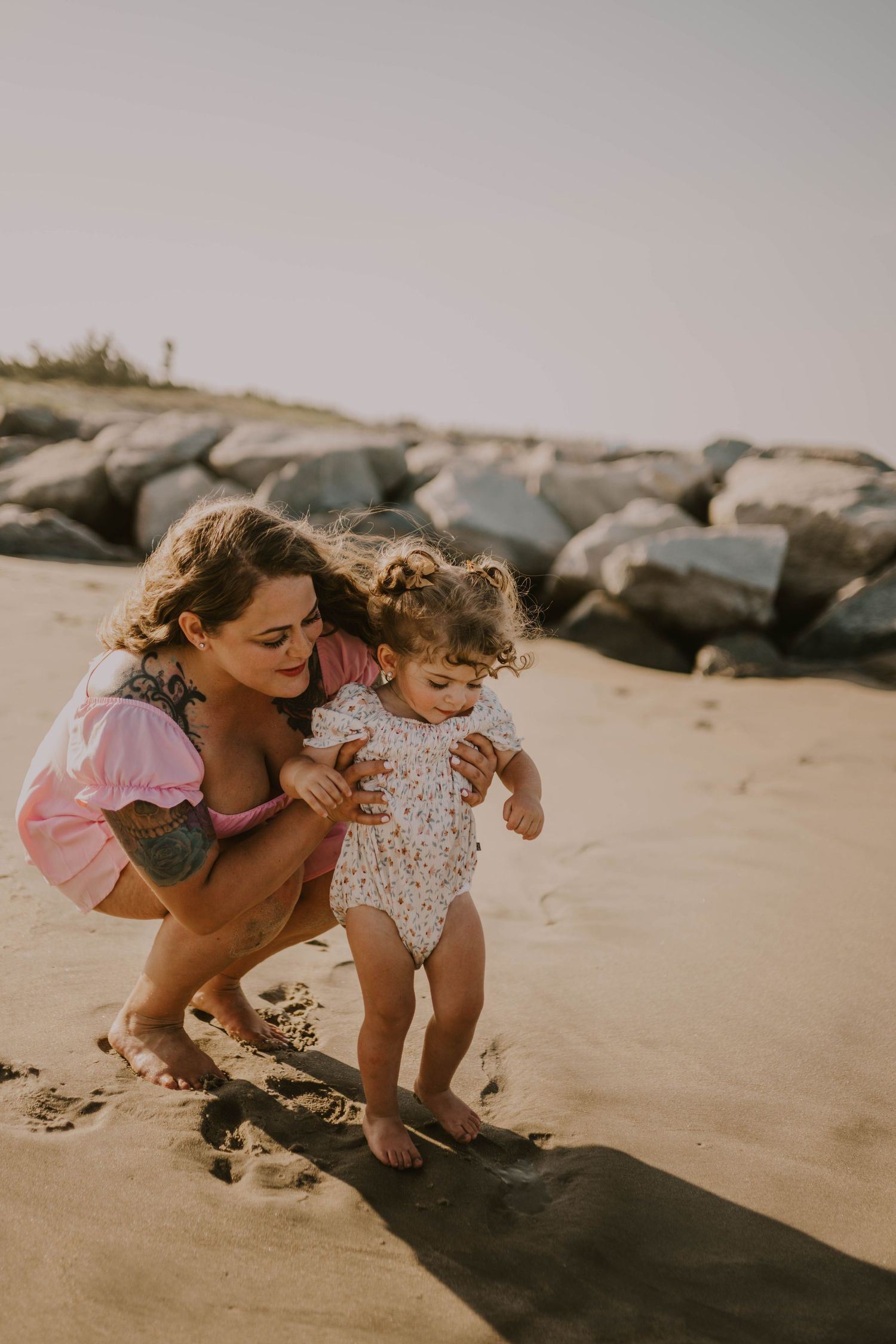 A mother crouches behind her toddler on a sandy beach, helping the child stand near large rocks.