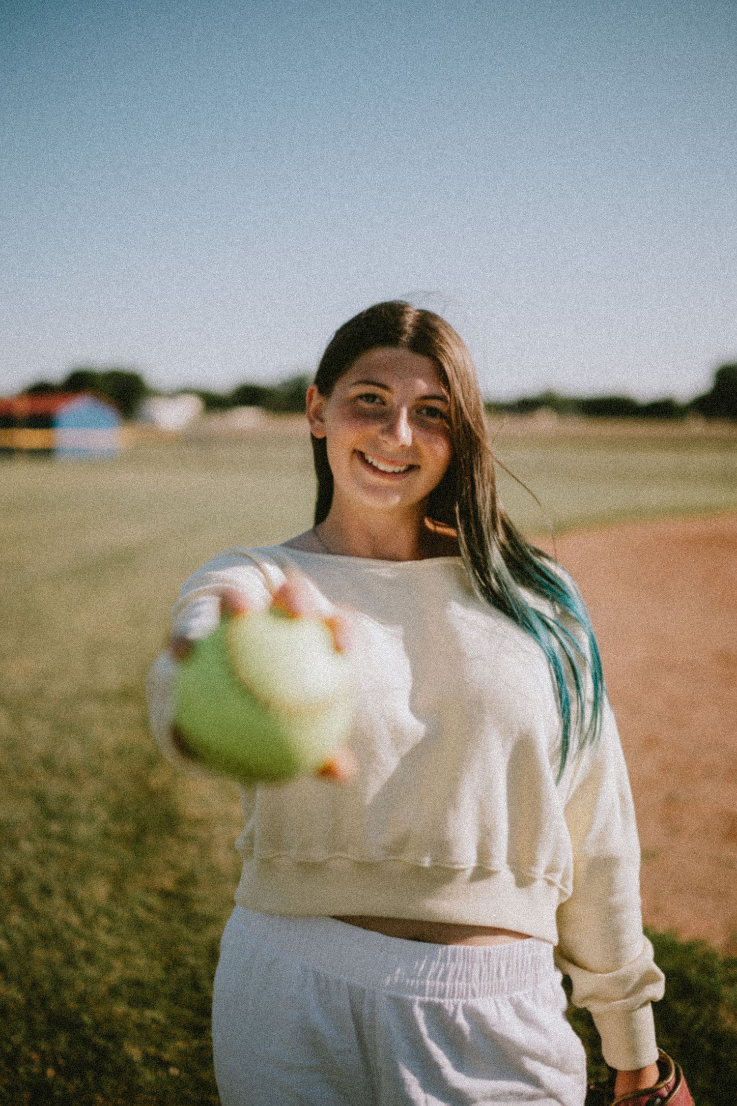 Smiling young woman with blue-tipped hair holding a softball on a sunny baseball field, wearing a white sweatsuit.