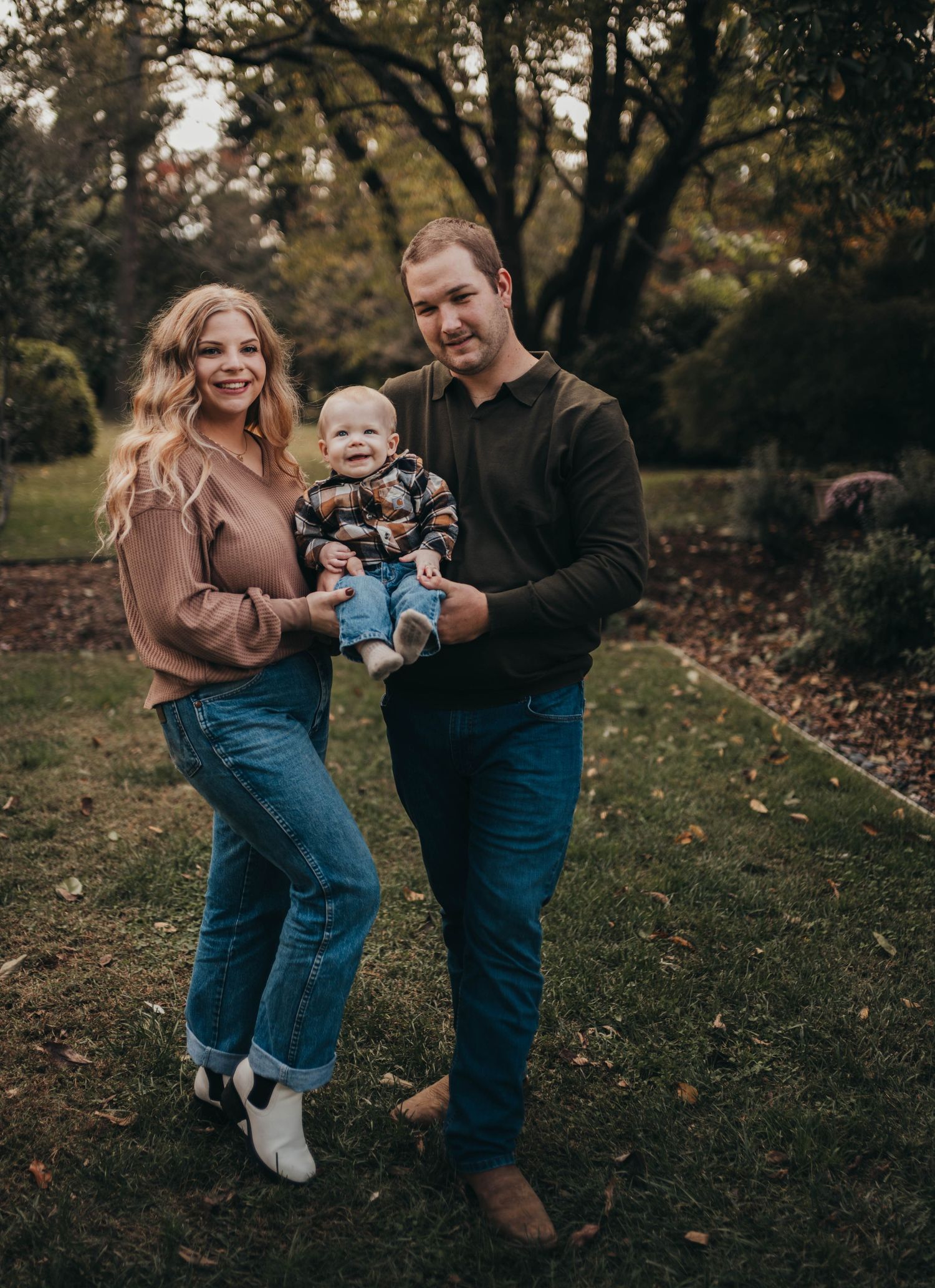 A family poses together outdoors during autumn with trees in the background on a grassy lawn.