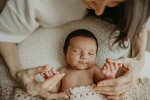 A sleeping newborn baby lies on a textured white blanket with hands held by someone in a white sweater. best newborn photographer