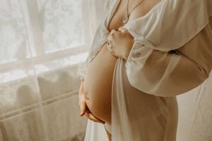 A pregnant woman in a white flowy dress stands by a window with soft natural light streaming through sheer curtains.
