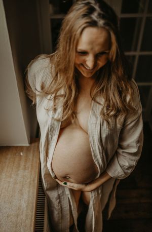 An intimate maternity portrait showing a pregnant woman in an open beige button-down shirt standing by a window.