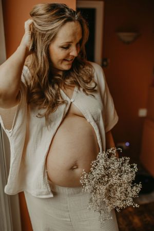 A pregnant woman in white clothing holds a delicate bouquet of baby's breath flowers in a warm-lit indoor setting.