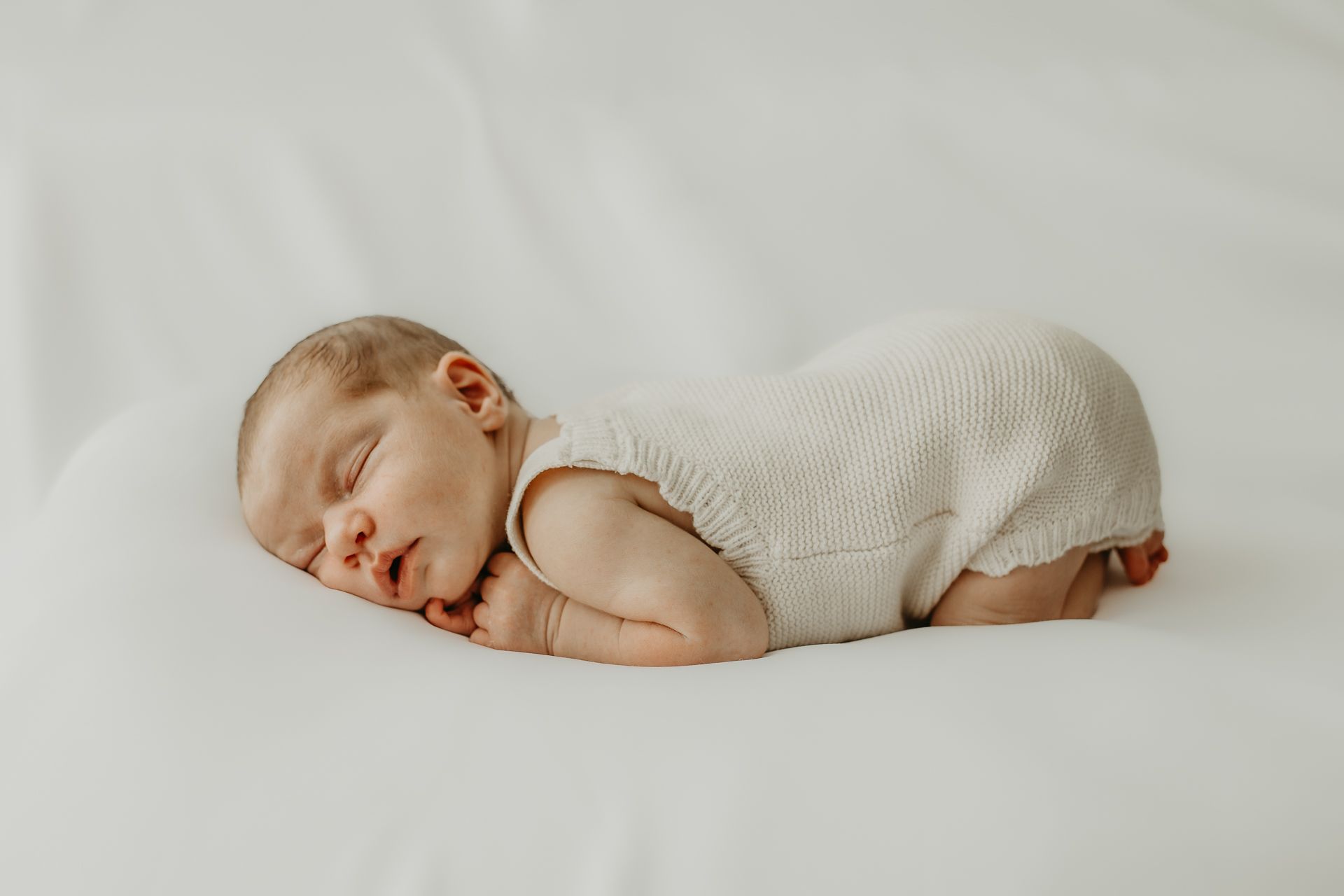 Peaceful sleeping newborn baby in white romper curled up on soft white background.