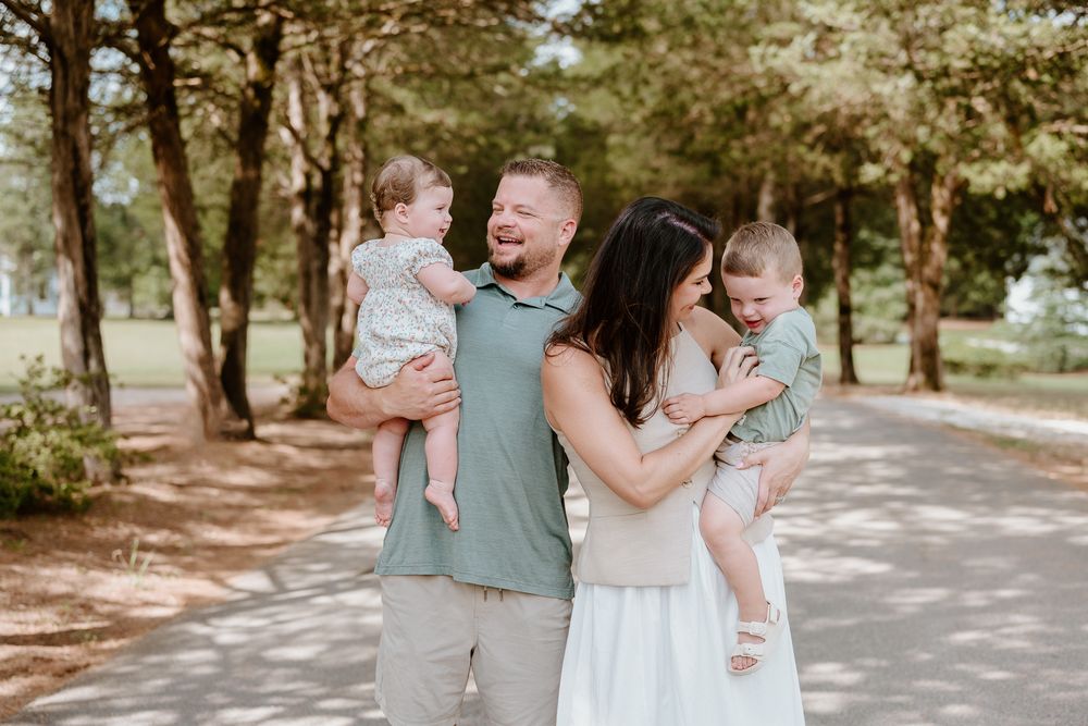 Family wearing coordinated blue and white outfits poses together on a tree-lined path in a park setting.