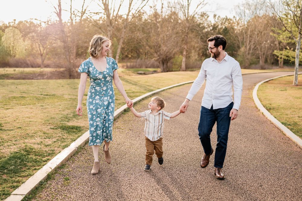 A family of 3 hold hands while walking together on a curved path in a golden-lit park setting during sunset.