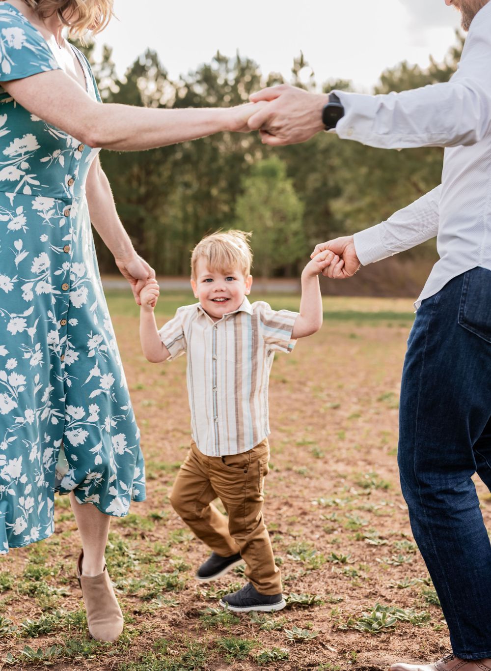 A young child in a striped shirt holds hands with adults in a blue dress and jeans while playing outdoors.