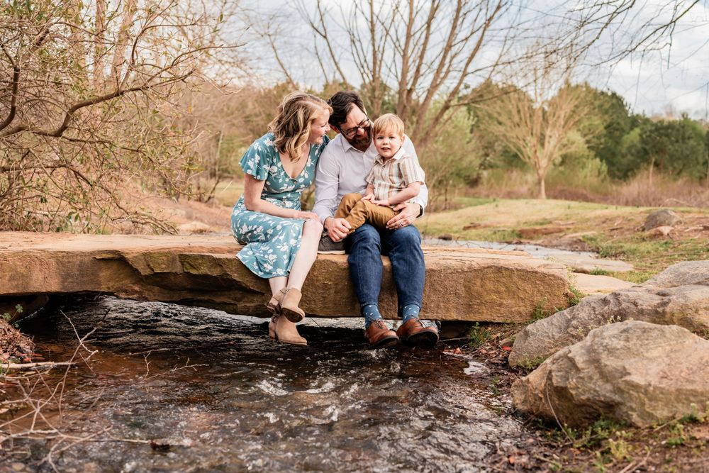 A family of 3 sit together on a large rock formation over a flowing stream in a natural outdoor setting.