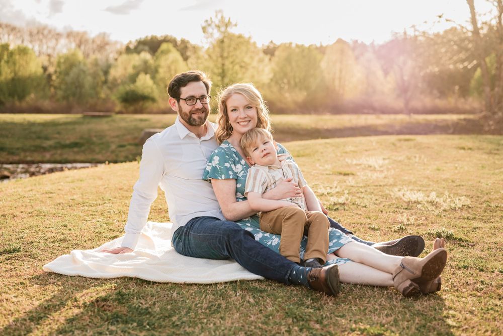 A family sits together on a white blanket in a sunlit meadow at golden hour holding blue flowers.