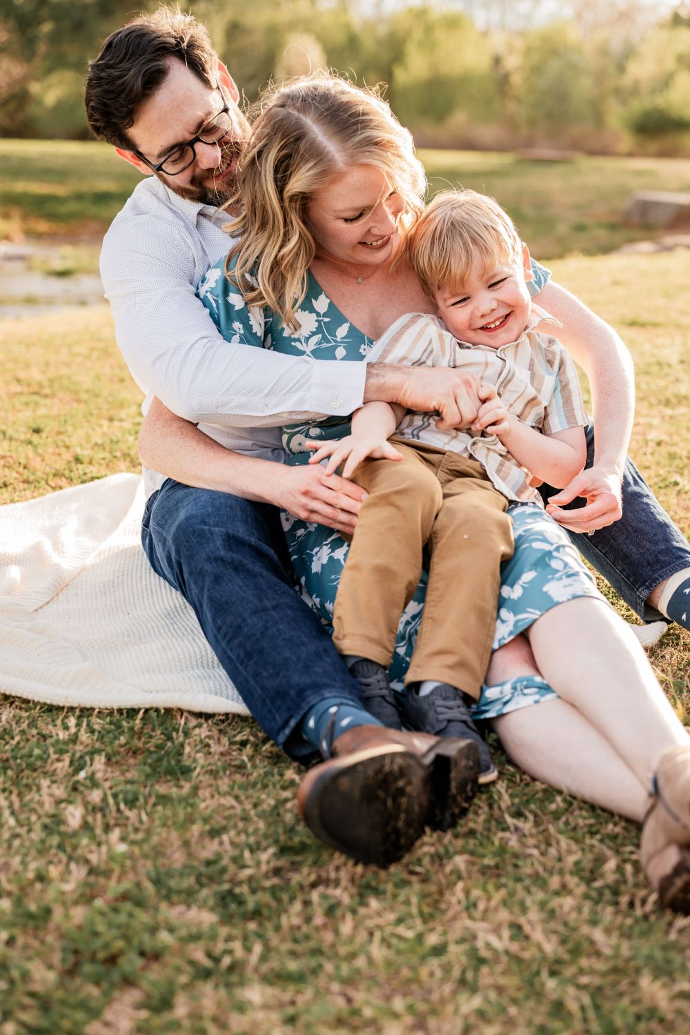 A family enjoys playful cuddles and laughter while sitting together on a blanket in a sun-lit park.