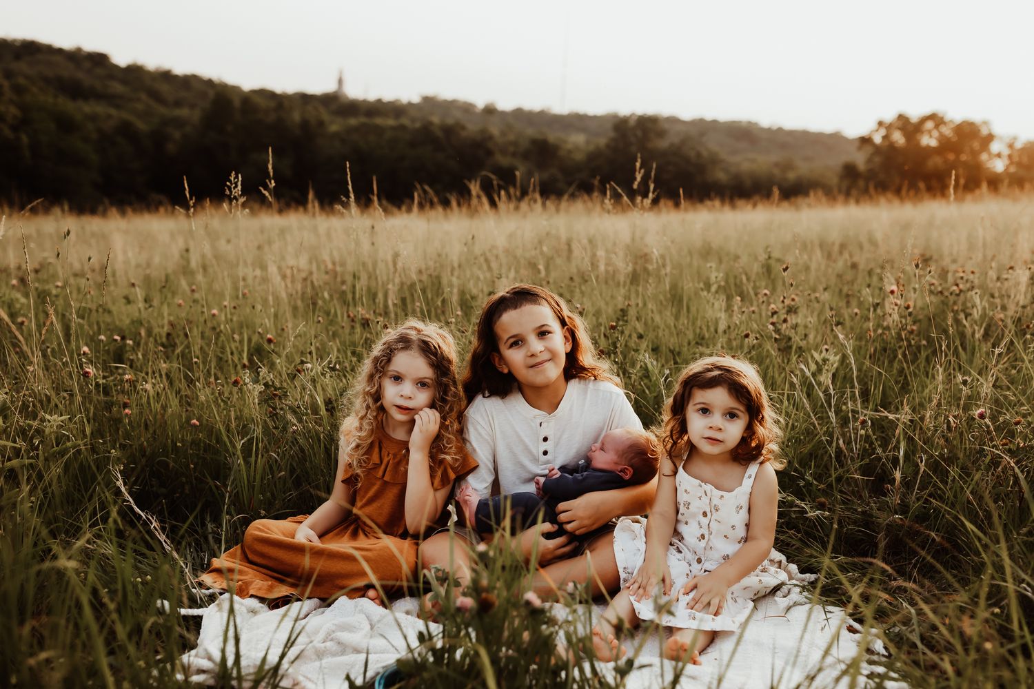 Three siblings sit together on a blanket in a golden field, the oldest child cradling a newborn baby.