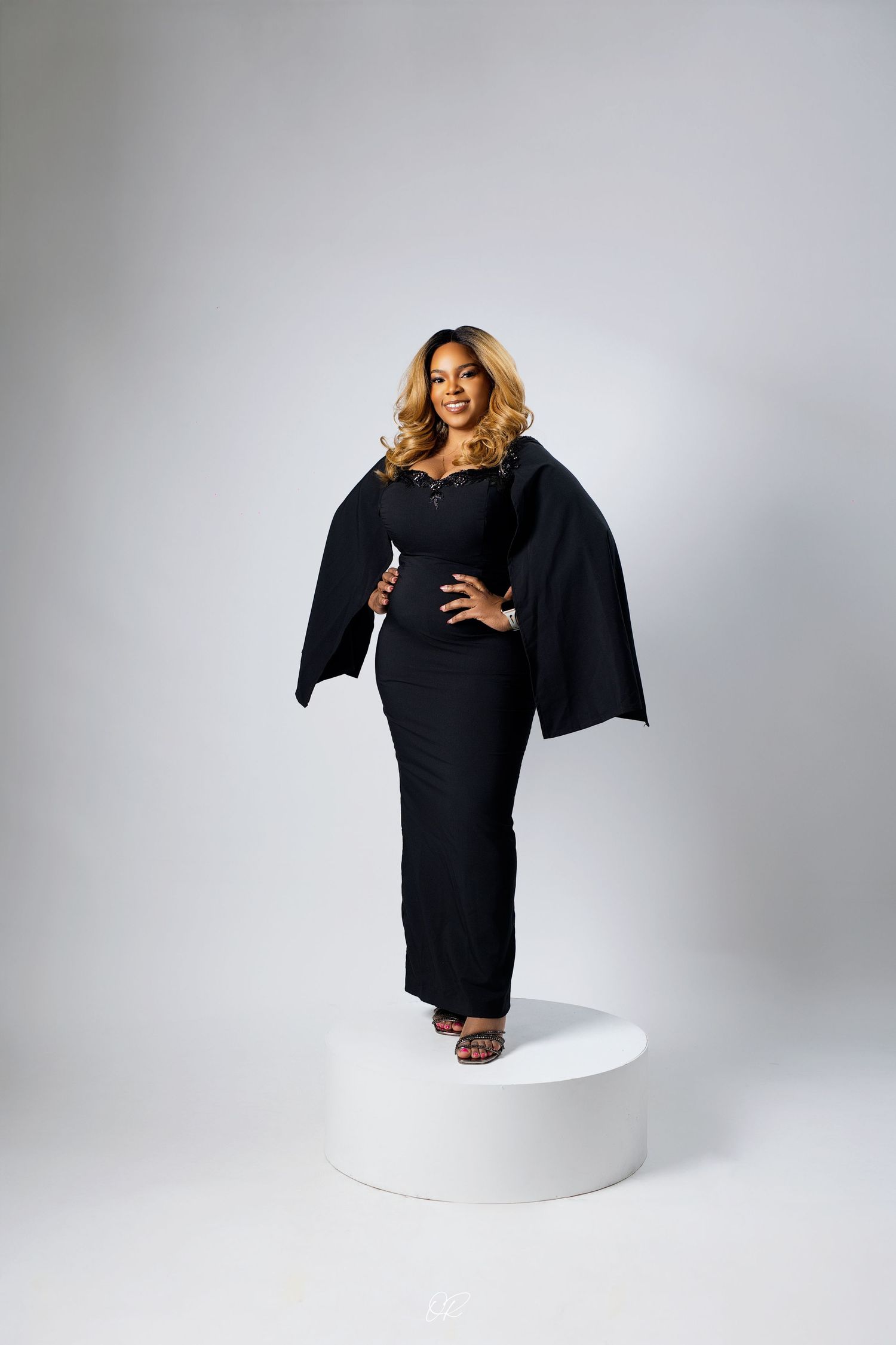 Elegant model poses in a black cape dress with dramatic sleeves on a white pedestal against light background.