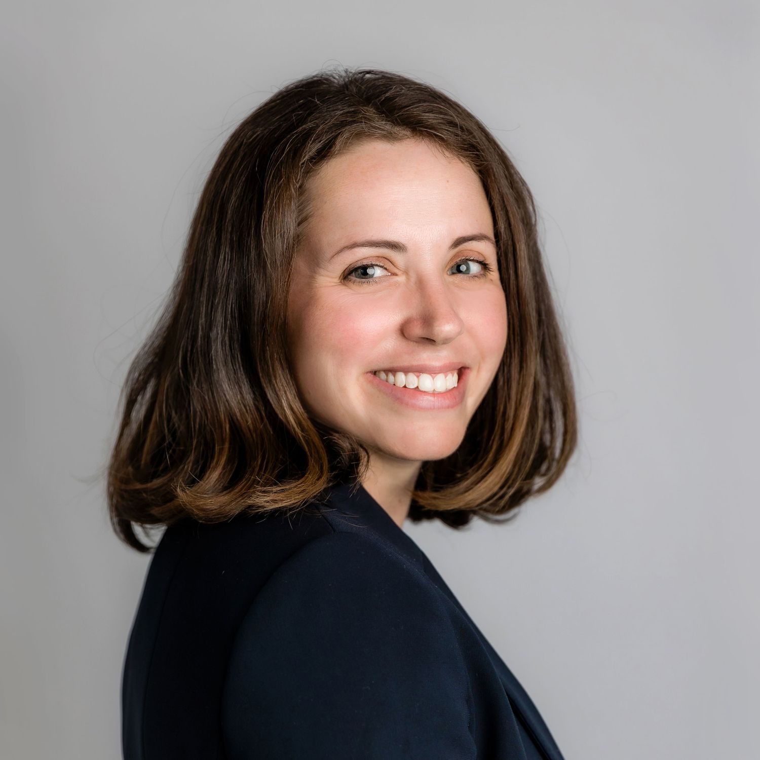 Warm and friendly Linkedin headshot of woman with medium-length brown hair wearing dark clothing against a light gray background