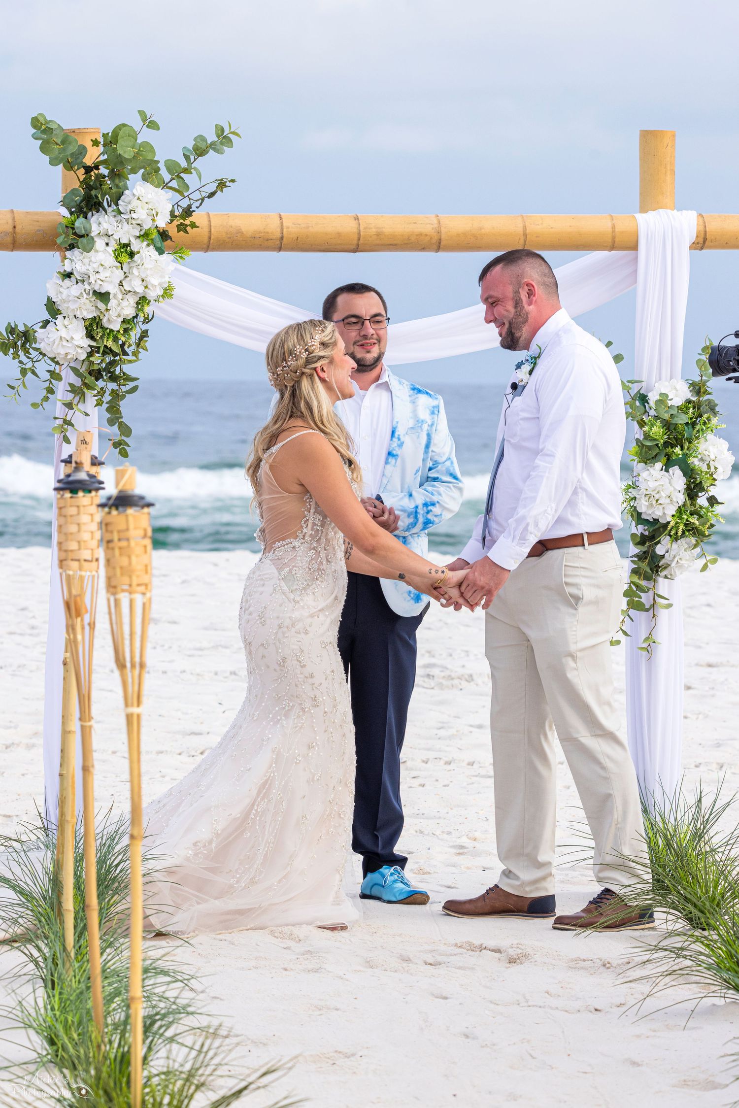 A beach wedding arch decorated with pampas grass and white fabric stands on a sandy shore near a dock.