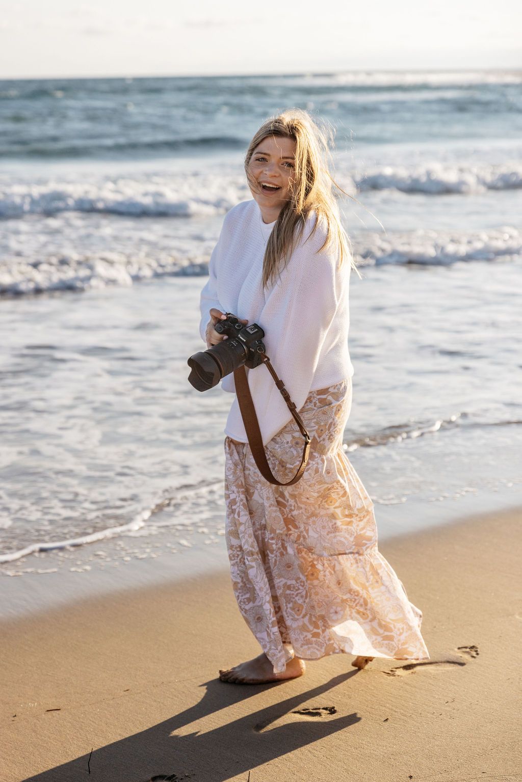 A smiling female photographer holding a camera while standing barefoot on a sunny beach.