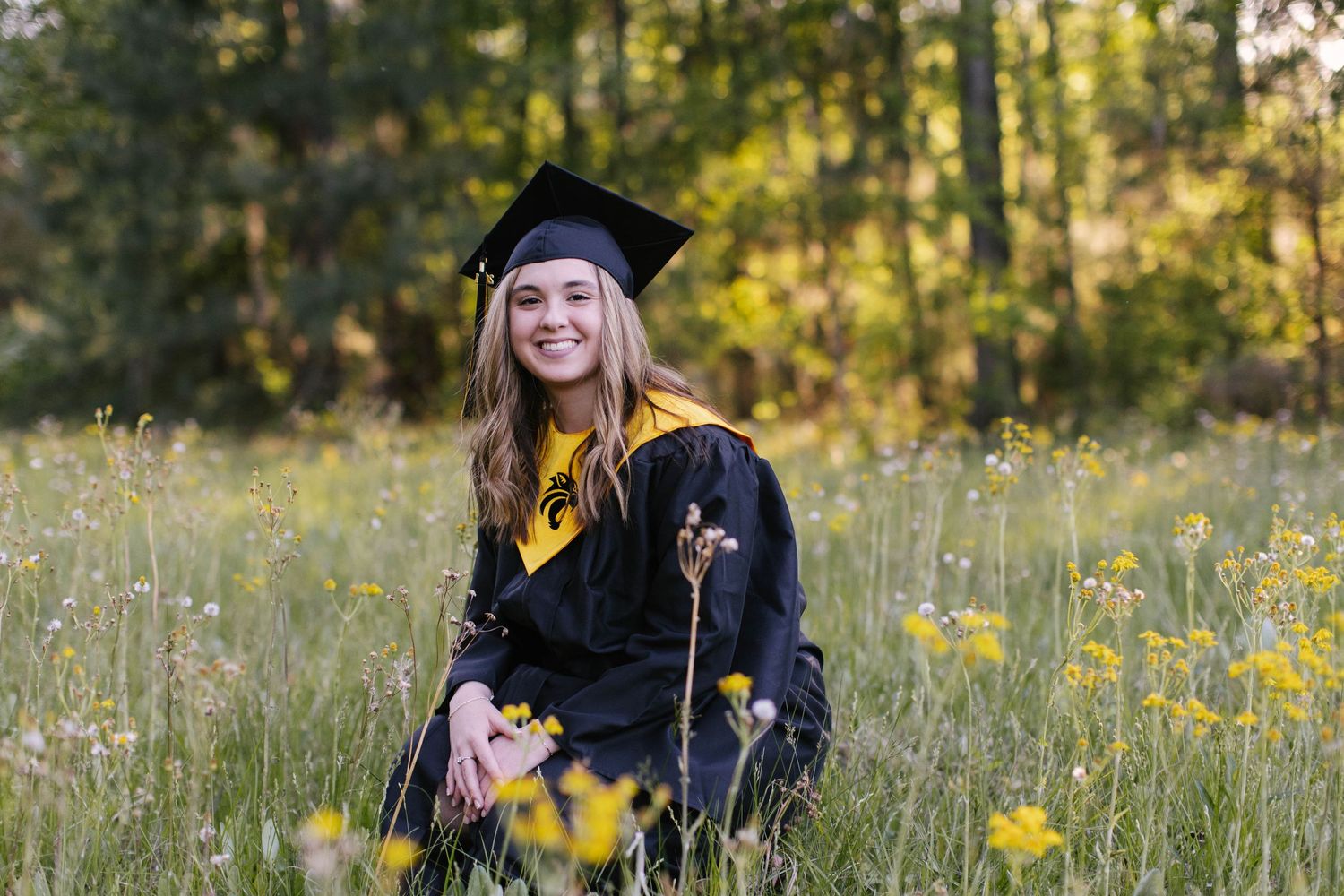 Graduate in cap and gown sitting in a field of yellow wildflowers during golden hour.