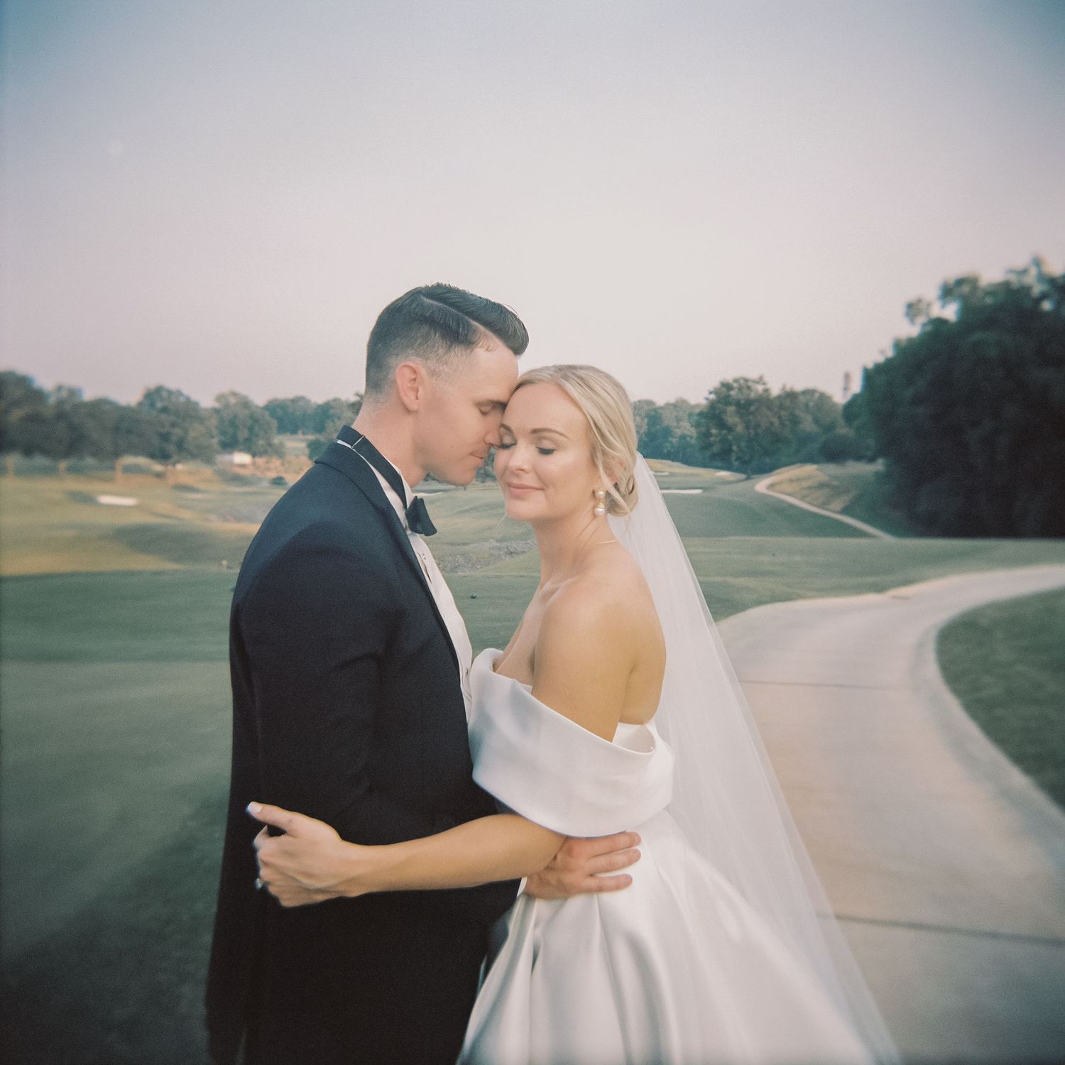 Bride and groom embracing at dusk on golf course, bride in off-shoulder gown with veil.