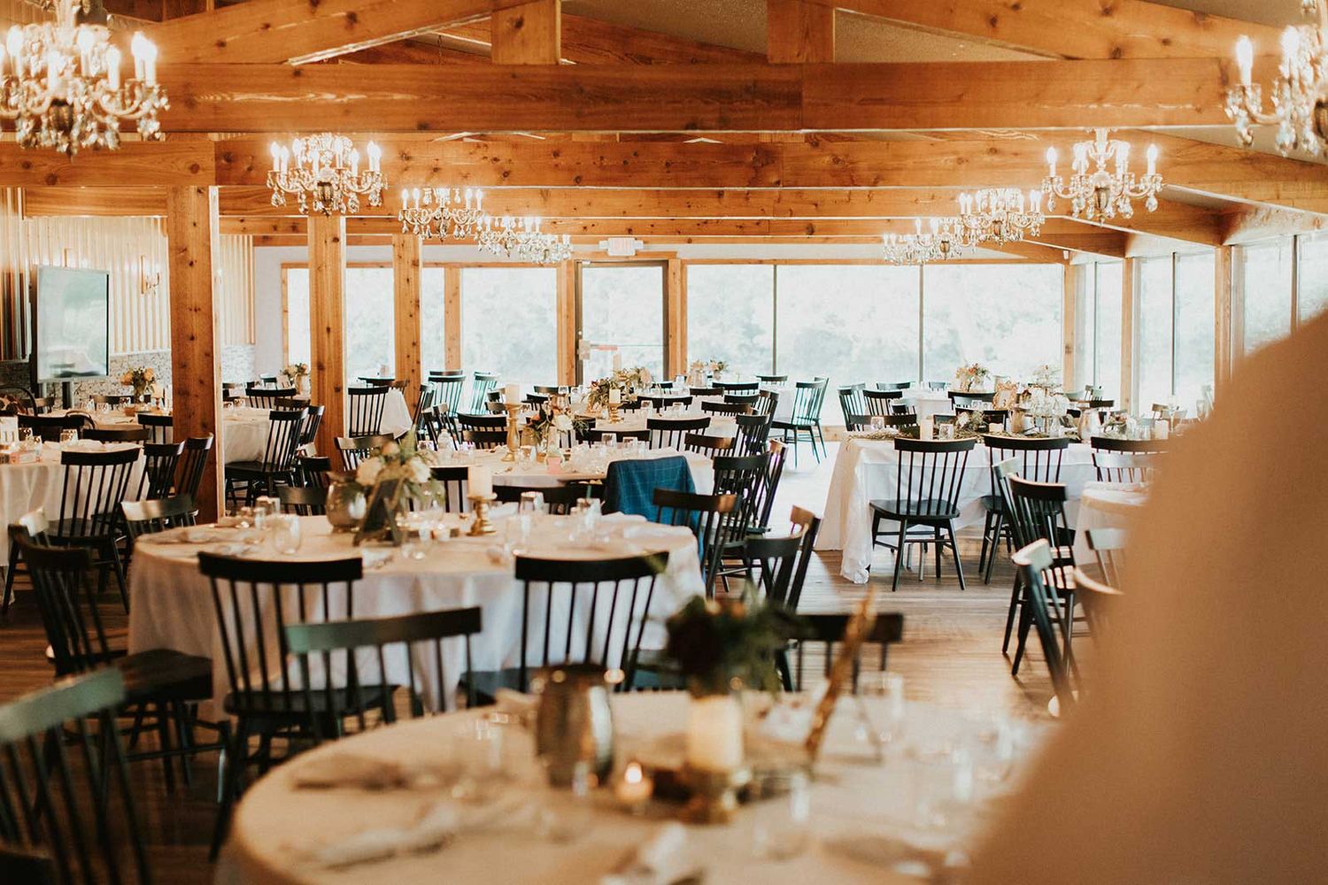 Spacious wedding venue interior with wooden beams, chandeliers, and round tables.