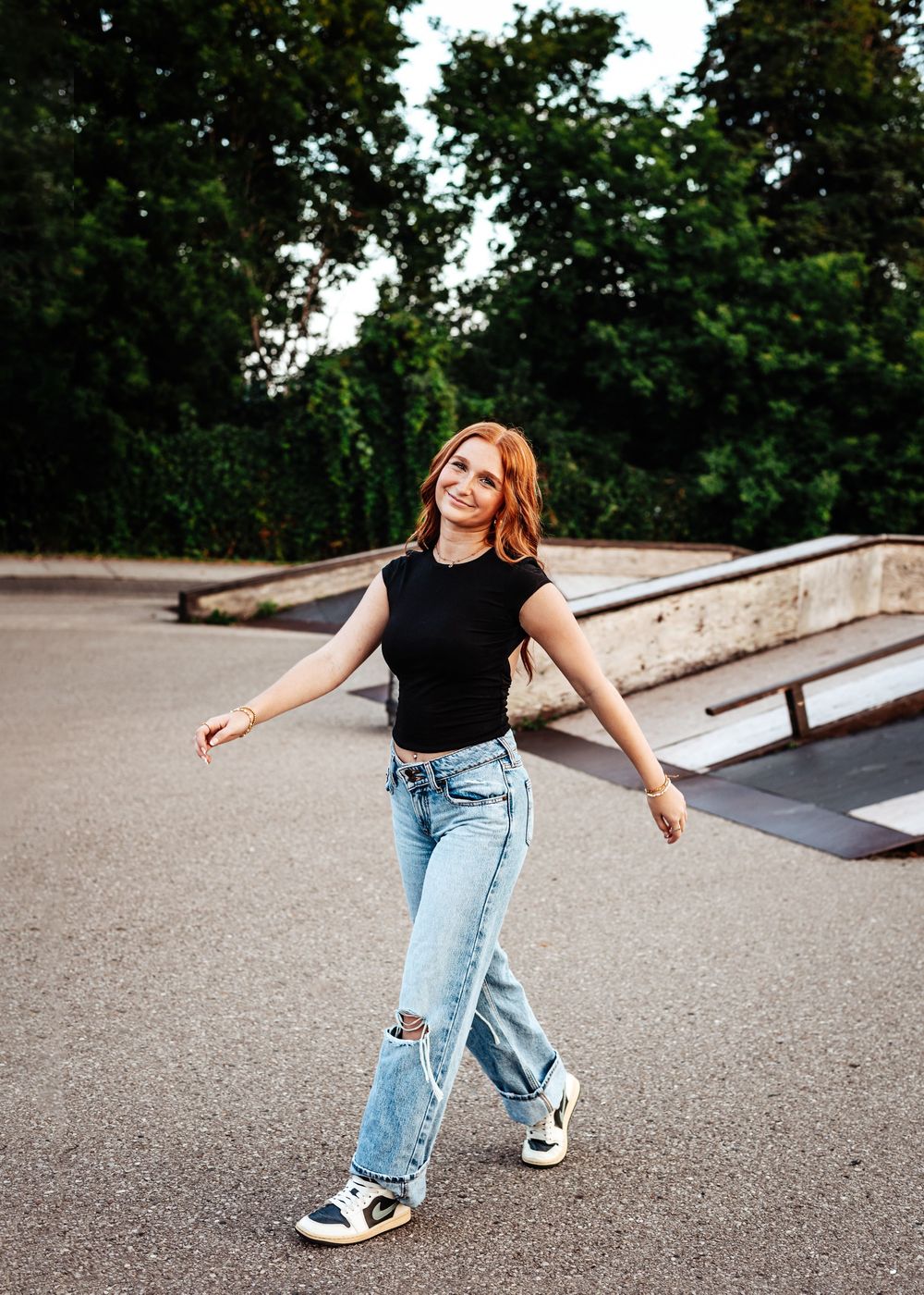 A person in a black t-shirt and ripped jeans poses playfully on a skate ramp with arms outstretched in summer.