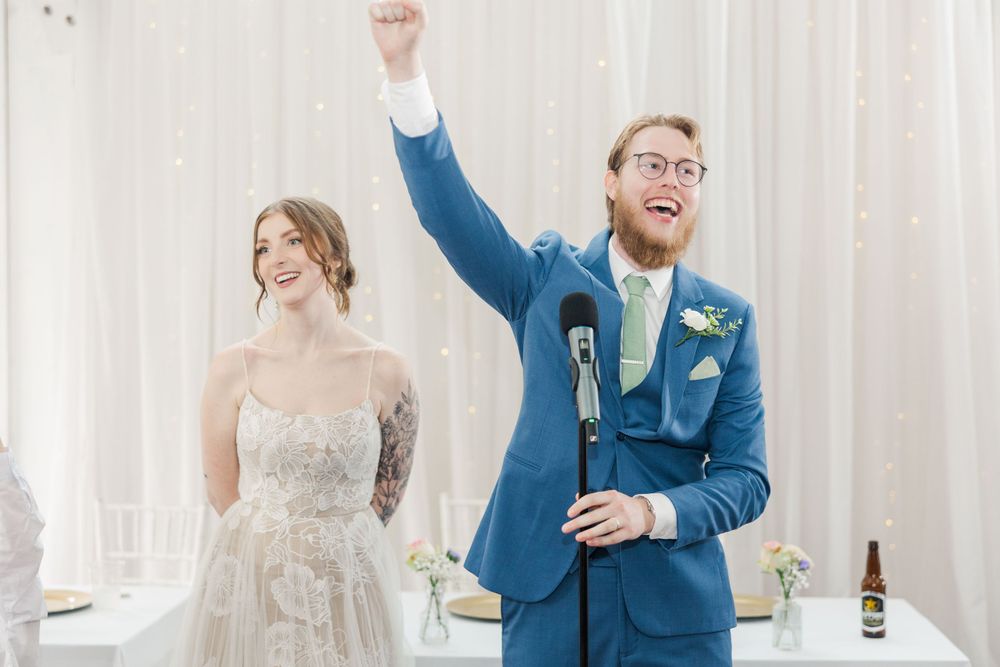 A couple celebrates during a wedding reception speech, with the groom raising his fist in excitement.