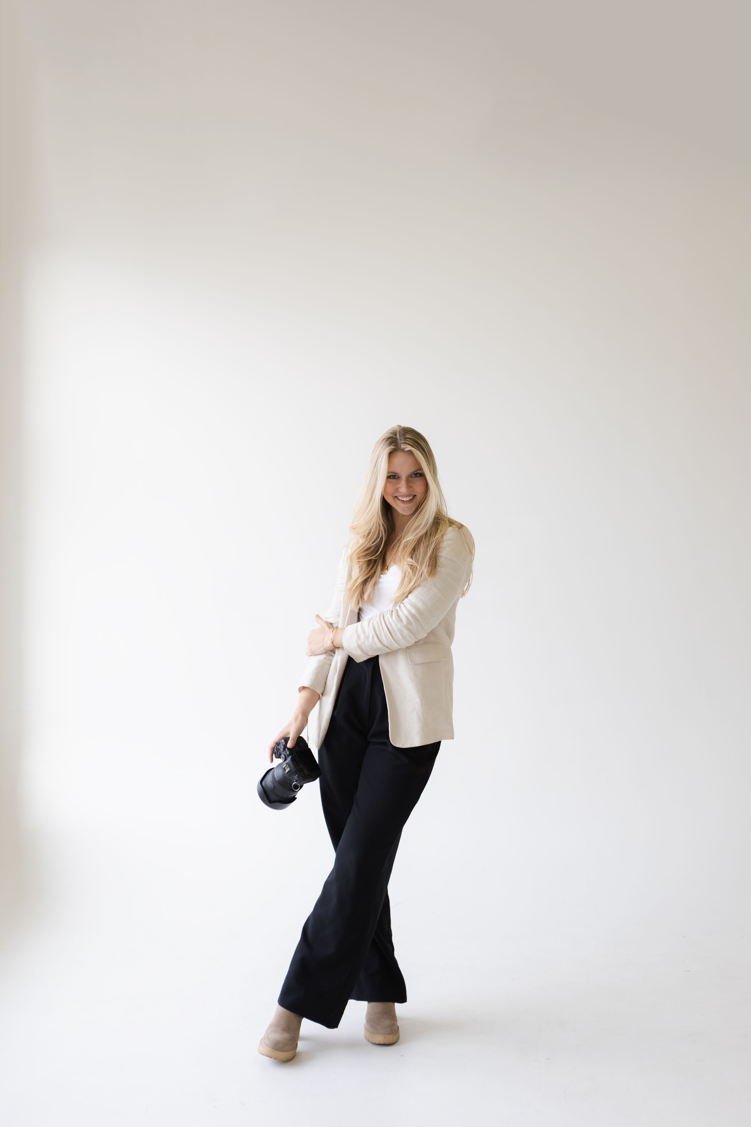 Woman in cream blazer and dark pants posing with camera against white background.