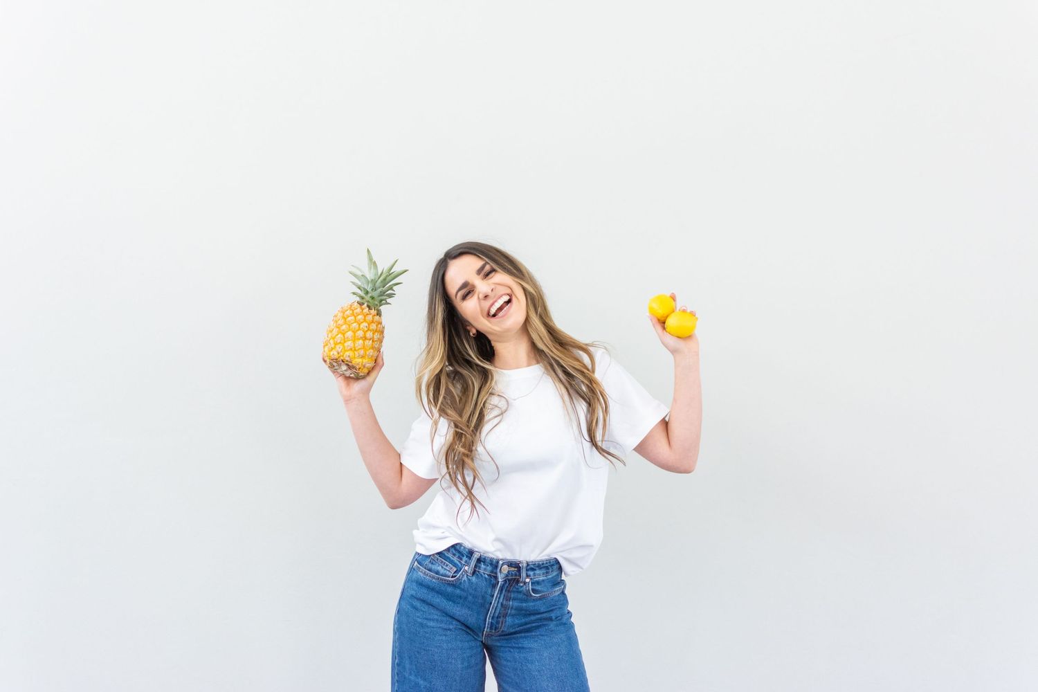 Woman in white t-shirt and jeans holds fresh pineapple and yellow lemon while smiling against white background.