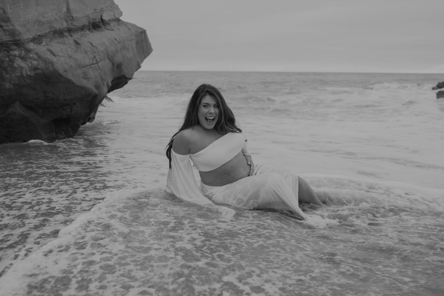 Black and white portrait of woman reclining in shallow ocean water near rocky shore.