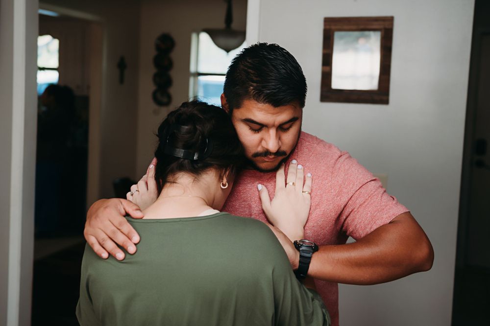 Two people share an intimate embrace in a dimly lit hallway, one wearing a salmon-colored t-shirt and another in olive green.