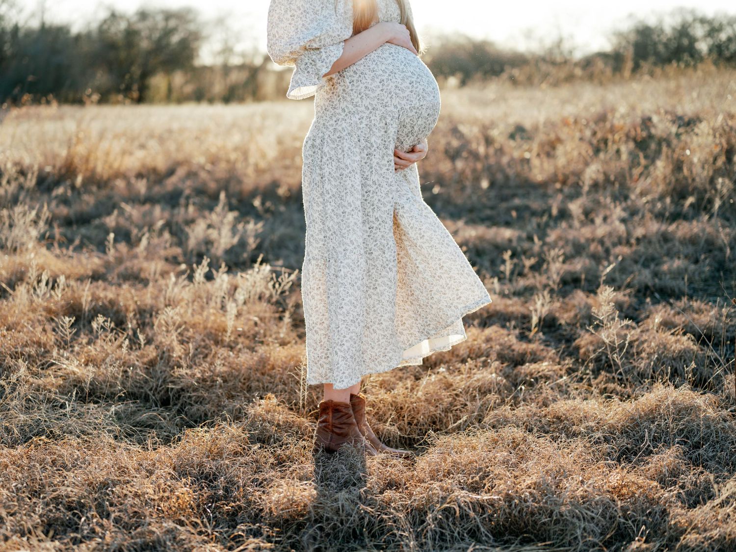 A flowing white polka dot dress sways in a sunlit field as brown boots stand among dried autumn grasses.