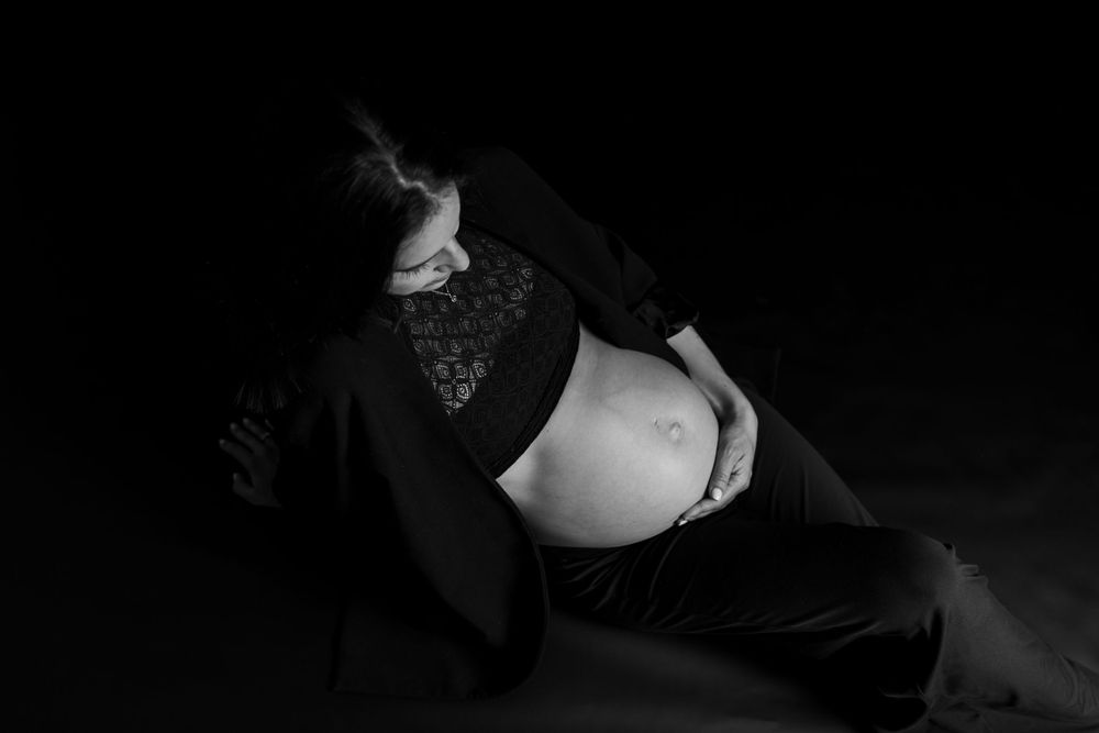 Ein dramatisches Schwarz-Weiß-Umstandsfoto zeigt eine werdende Mutter, die in dunkler Kleidung vor einem dunklen Hintergrund liegt.