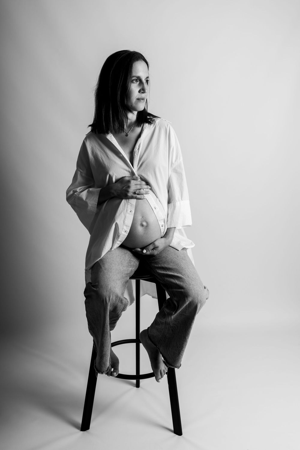 Schwarz-weißes Schwangerschaftsfoto einer Frau in weißem Hemd und Hose, die mit freiliegendem schwangeren Bauch auf einem Hocker sitzt.