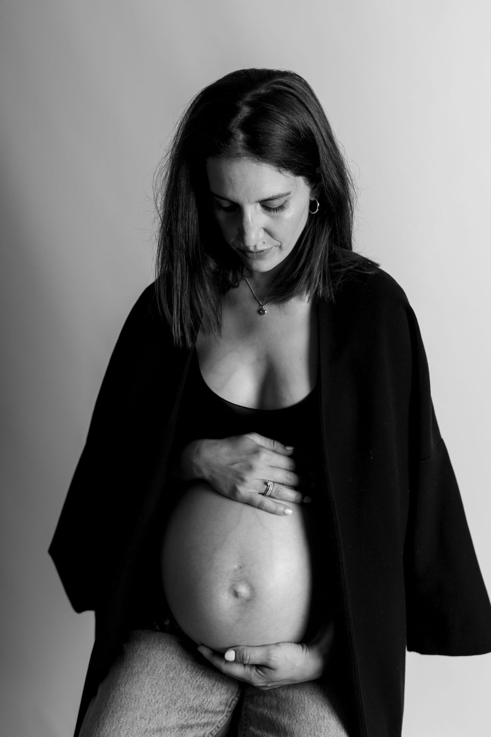 Schwarz-weißes Schwangerschaftsfoto einer werdenden Mutter, die einen dunklen Blazer trägt und auf ihren schwangeren Bauch blickt.