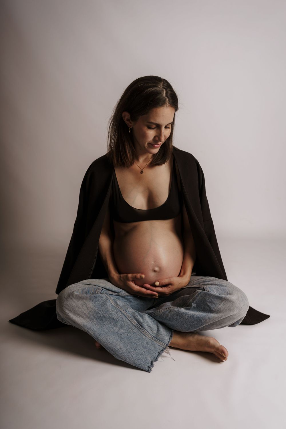 Eine Frau in schwarzer Loungewear sitzt im Schneidersitz und wiegt ihren schwangeren Bauch vor einem minimalistischen, neutralen Hintergrun