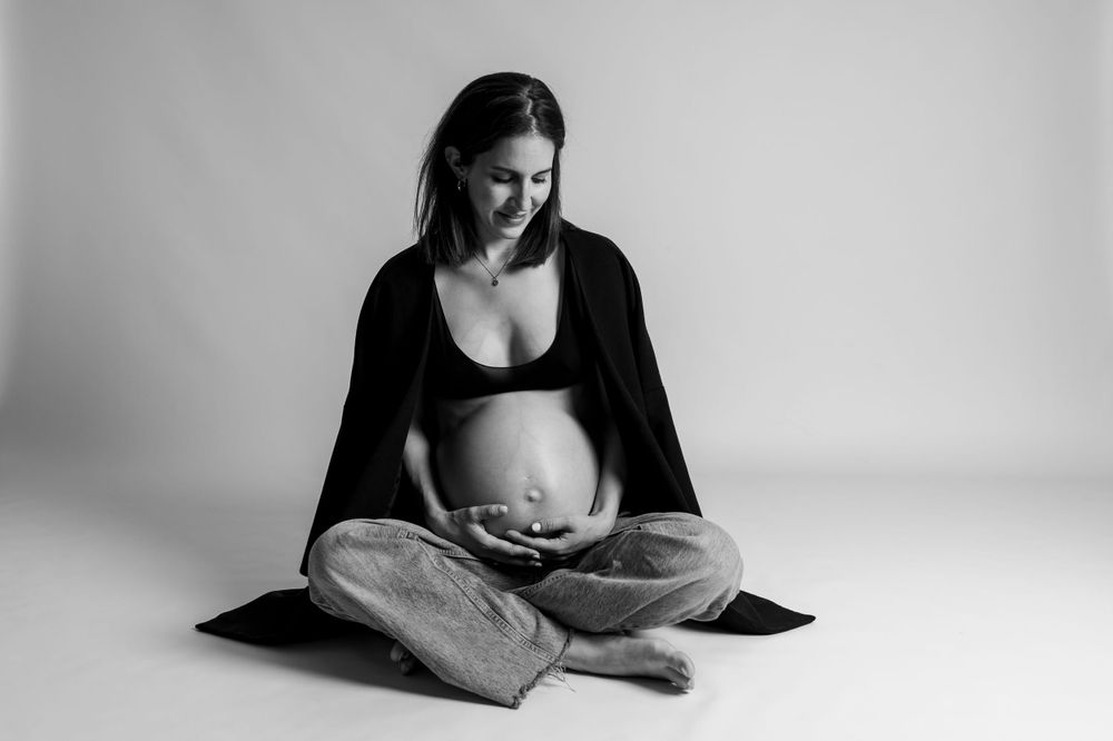 Schwarz-Weiß-Foto einer schwangeren Frau, die mit gekreuzten Beinen auf dem Boden sitzt, einen Blazer trägt und ihren Babybauch hält.