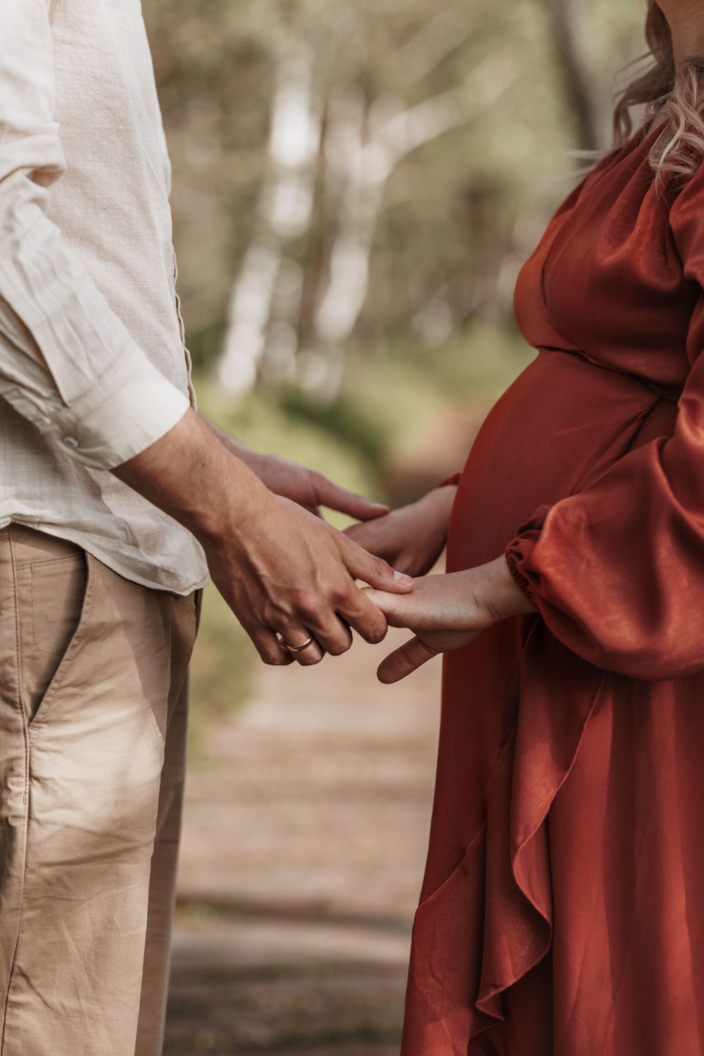 Eine schwangere Frau in einem fließenden roten Kleid hält Händchen mit ihrem Partner vor einem verschwommenen Naturhintergrund.