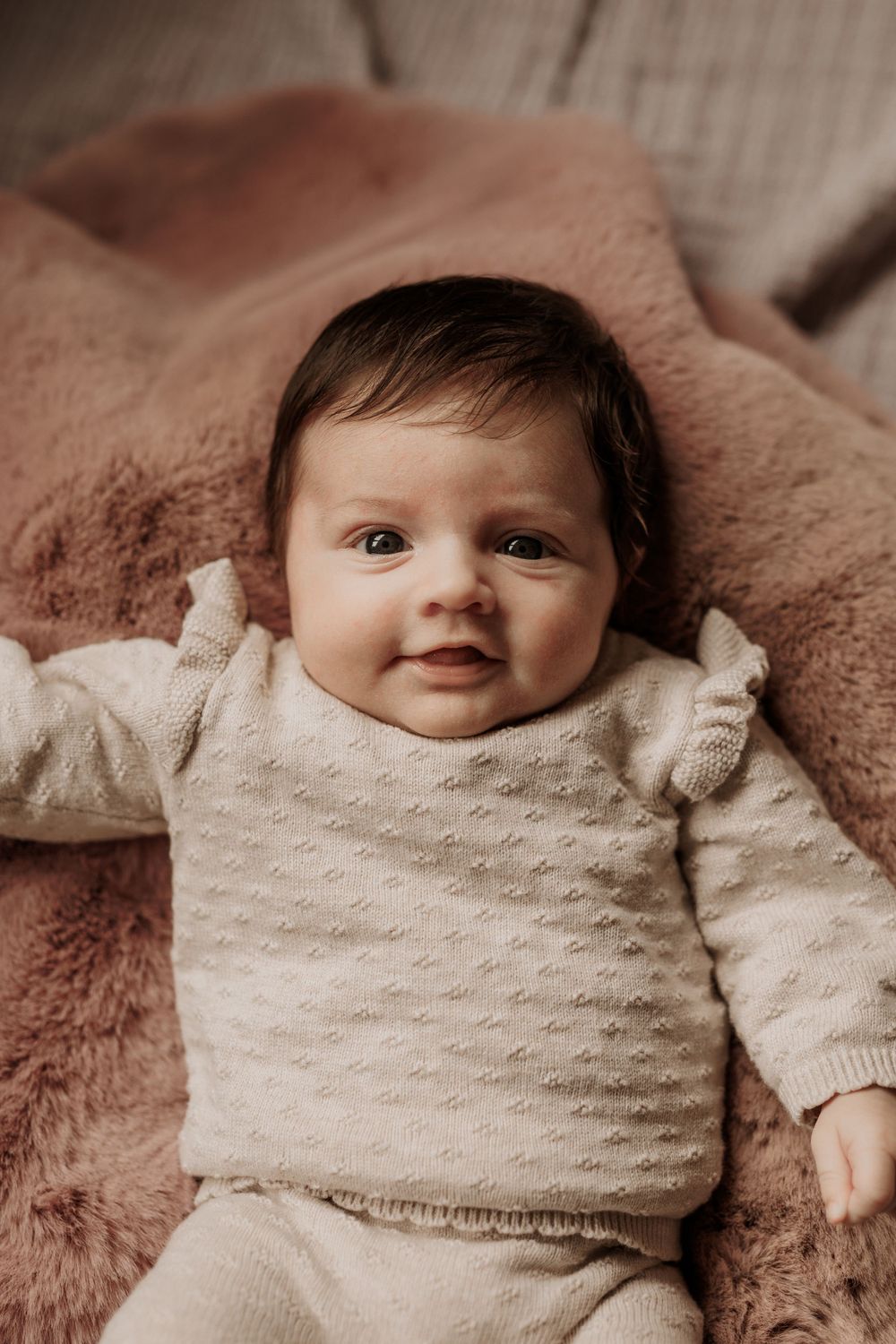 Ein Baby in einem kuscheligen cremefarbenen Pullover liegt auf einer zartrosa Decke und lächelt süß.