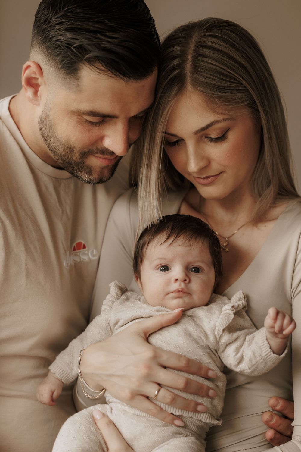 Intimate family portrait in warm neutral tones showing parents embracing their baby in matching beige outfits.