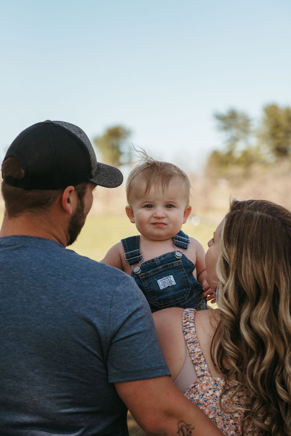Family moment captured outdoors with baby in overalls against a sunny backdrop.