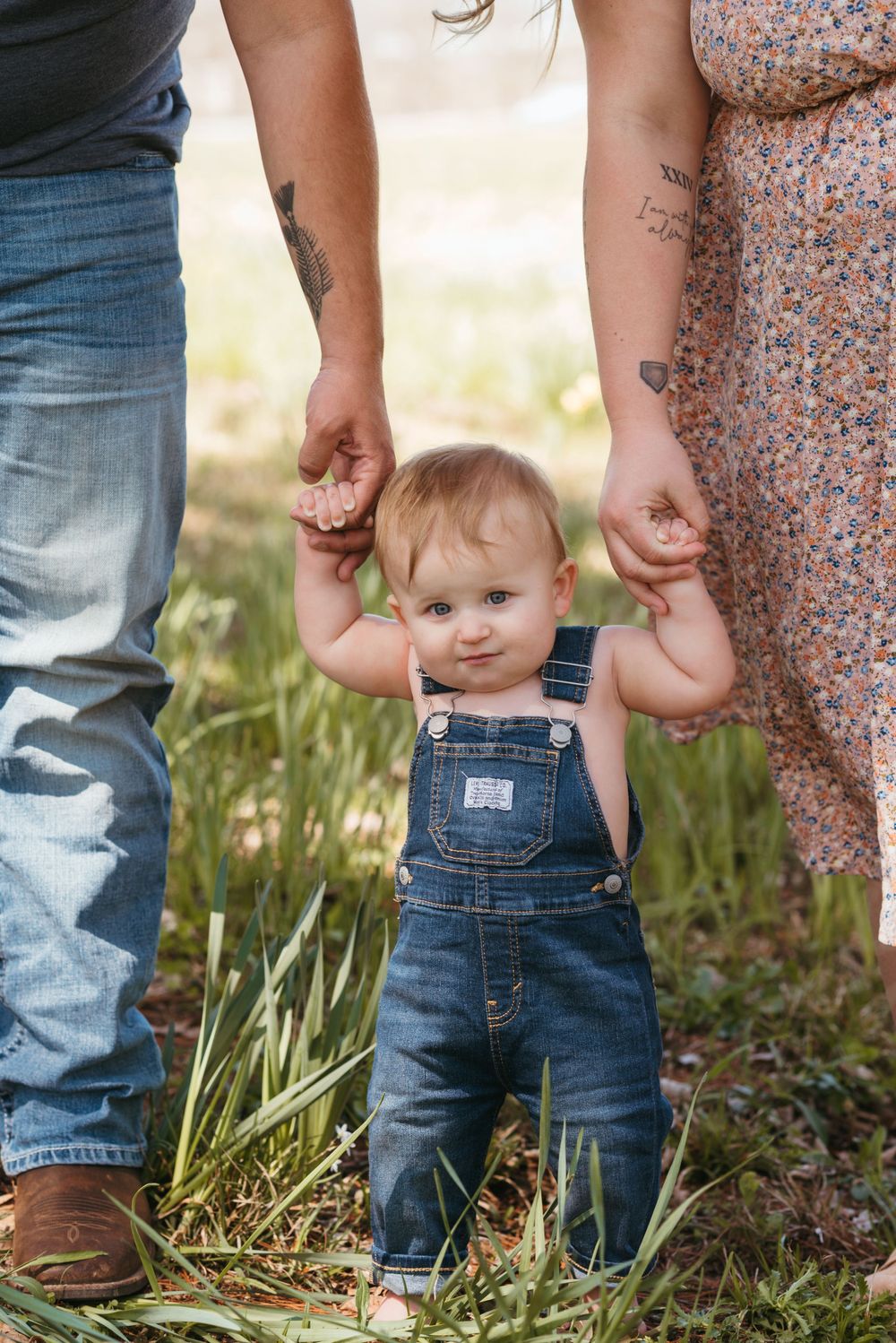 Baby in denim overalls takes first steps while holding parents' hands in a grassy field.