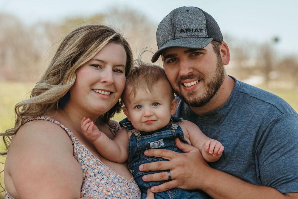 Family poses together outdoors with baby wearing denim overalls in sunny field.