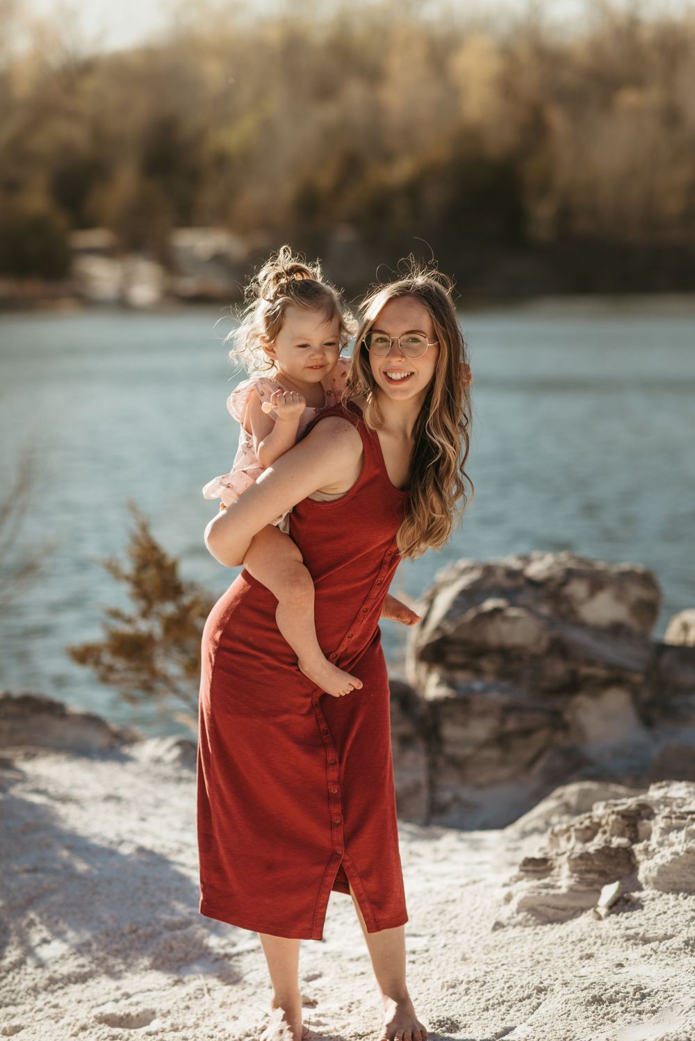 A parent and child share a joyful moment by the water, wearing a flowing red dress.