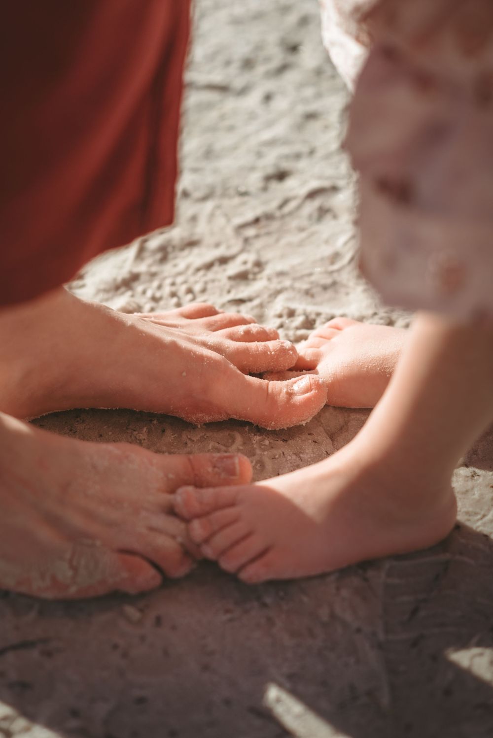 Close-up of adult and child feet touching on sandy ground in warm sunlight.